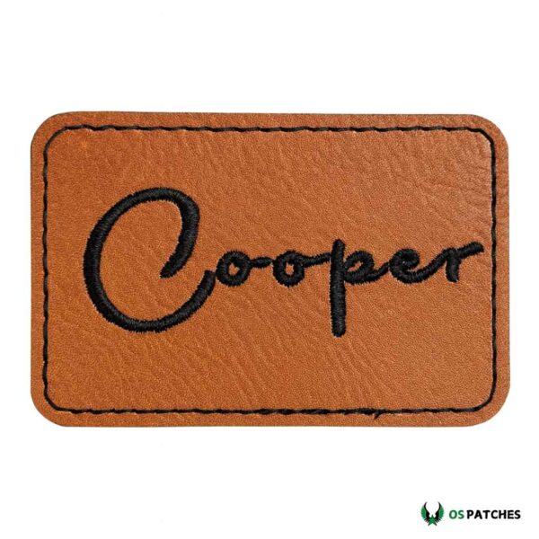 Rectangular brown leather patch with embroidered black text “Cooper.”