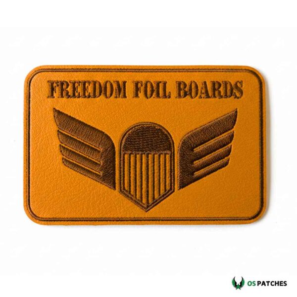 Rectangular tan leather embroidered patch with text "Freedom Foil Boards" and winged shield design.