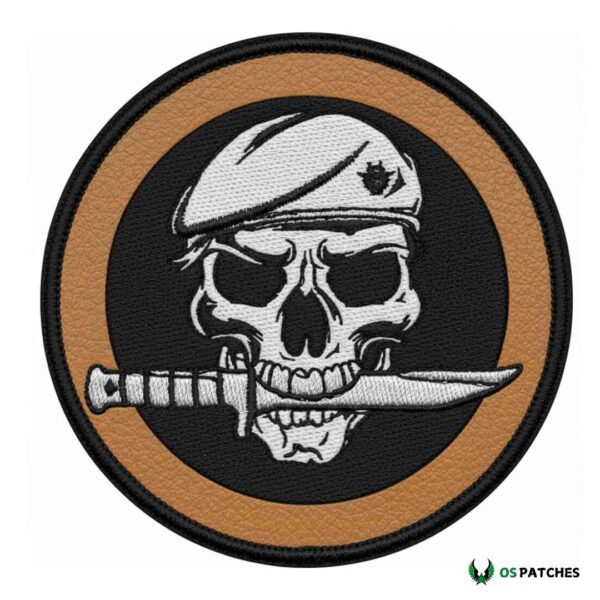 Round brown leather embroidered patch with skull wearing beret and holding knife in mouth.